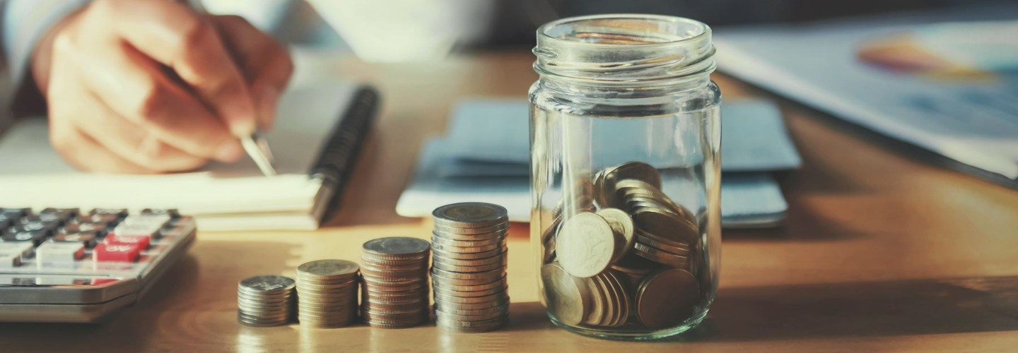 a jar of coins next to a stack of coins, calculator and a hand writing with a p;en