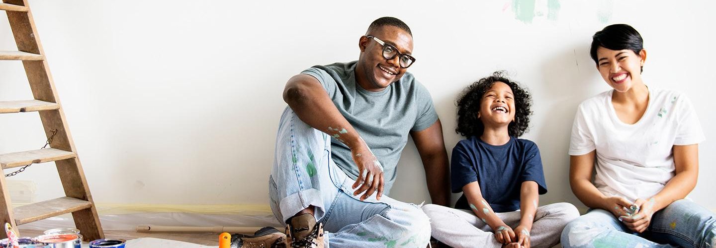 A family of three smiling sitting against a wall in their house